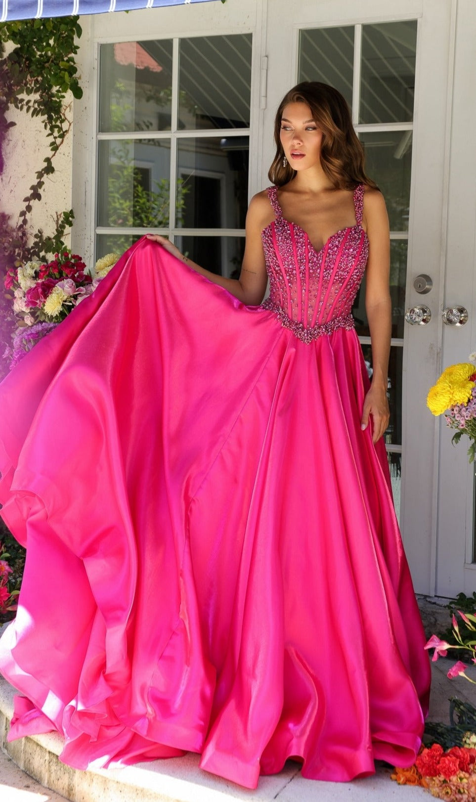 A woman stands outside in front of a glass door, surrounded by flowers, holding the skirt of the Long Formal Dress 29521 by Ava Presley—a bright pink ball gown with a beaded corset bodice.